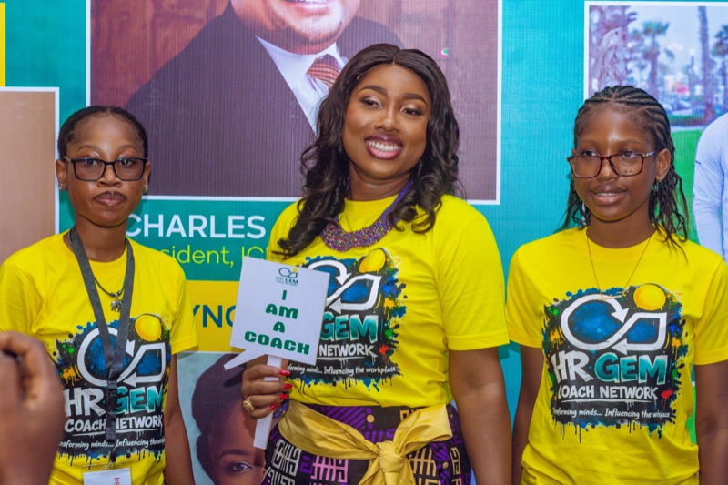 Three team members in yellow HR GEMs T-shirts at the speakers wall, one holding an "I am a coach" placard