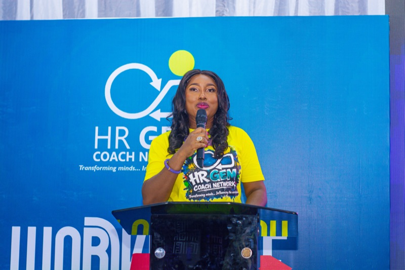 A host in a yellow HR GEMs T-shirt speaking at the podium
